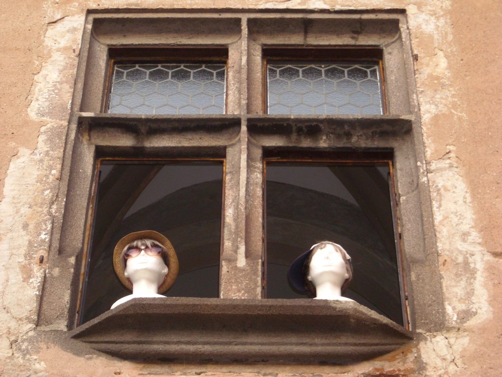 V okne sú dve figurínové hlavy. Jedna má klobúk a slnečné okuliare a druhá šiltovku. Vyzerajú akoby sledovali okolie.