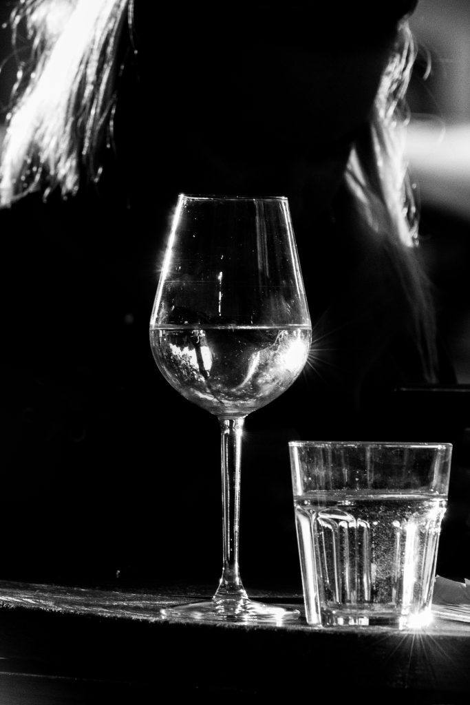 ČB fotografia, v pozadí je žena, vidno ju veľmi slabo. Pred ňou je vidno pohár na víno a vodu.