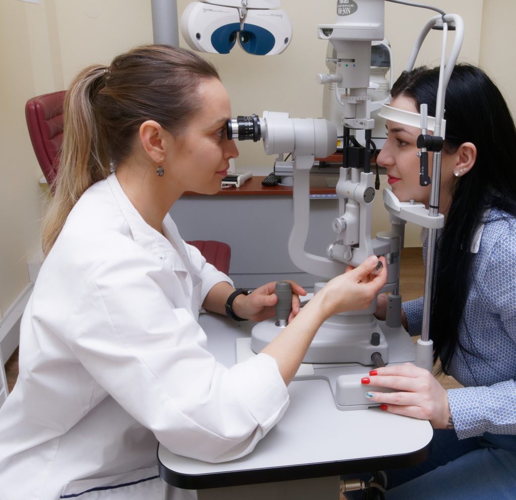 Na obrázku je lekárka, ktorá pozerá do prístroja na očné vyšetrenie. Oproti nej sedí pacientka s hlavou položenou na meracom prístroji. 