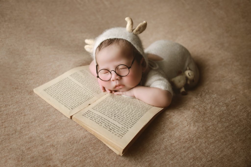 Na štylizovanej fotografii je spiace bábätko v okuliarkoch a hlávku má položenú na otvorenej knihe. 