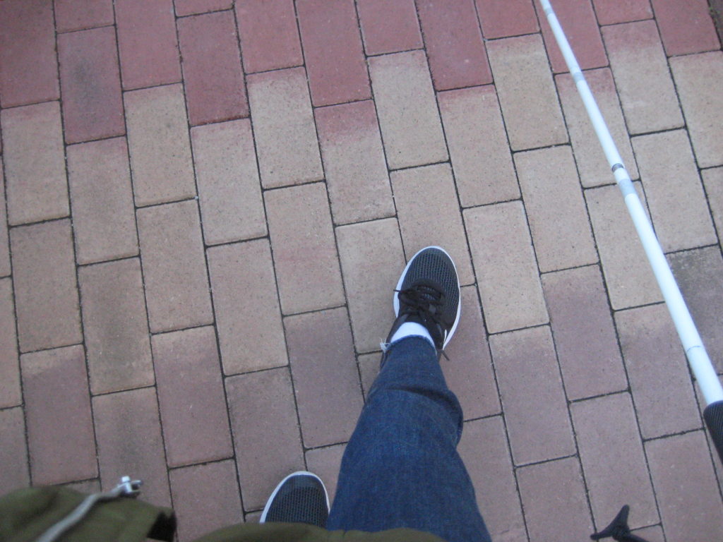 Na obrázku je záber na kráčajúceho človeka, ktorému vidno len nohy a bielu palicu. 