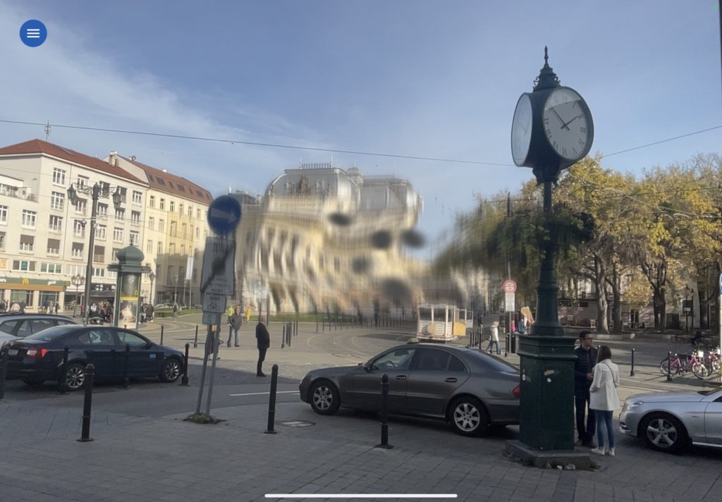 Na fotografii je záber z Hviezdoslavovho námestia v Bratislave. Správne by mal uprostred fotografie človek vidieť historickú budovu SND, avšak je celá pokrútená a rozmazaná. Po stranách vidno iné budovy, hodiny, autá a ľudí na námestí.