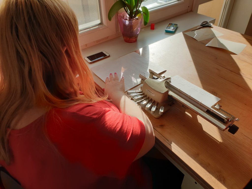 Žena, ktorú vidno od chrba, sedí za stolom, na ňom ma Pichtov stroj a rukami číta materiál v Braillovom písme.