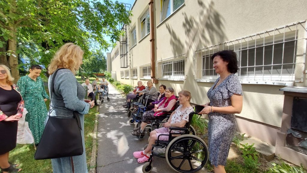 Na obrázku sú pred budovou seniori sediaci vo vozíkoch. Vedľa nich žena, zrejme pracovníčka zariadenia a oproti nim Z. Stavrovská v spoločnom rozhovore.