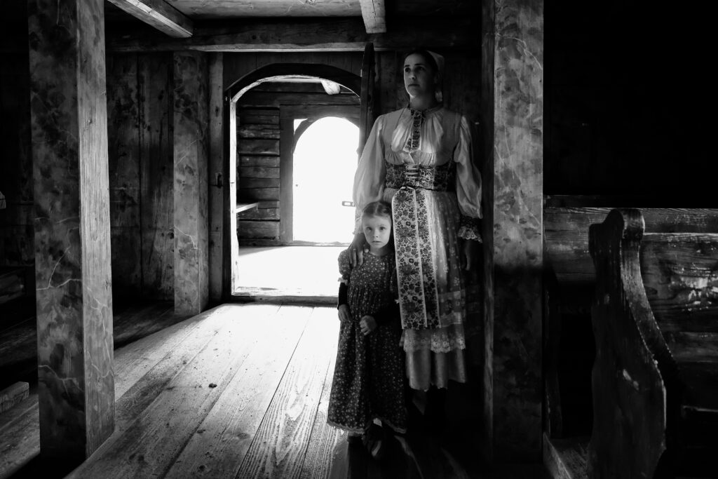 Na obrázku je žena oblečená v kroji, pri nej stojí dievčatko, tiež oblečené v dobových šatách. Stoja v priestoroch dreveného kostola.