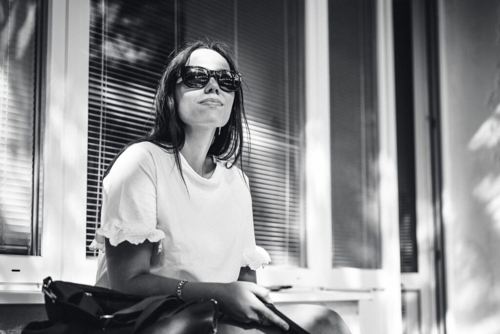 Na ČB foto je portrét mladej ženy sediacej na lavičke pred oknami. Na tvári má slnečné okuliare. 