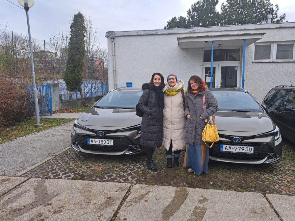 Autá sú odparkované na parkovisku ÚNSS a medzi nimi stoja smiate T. Winterová, S. Tóthová a Z. Egydová.